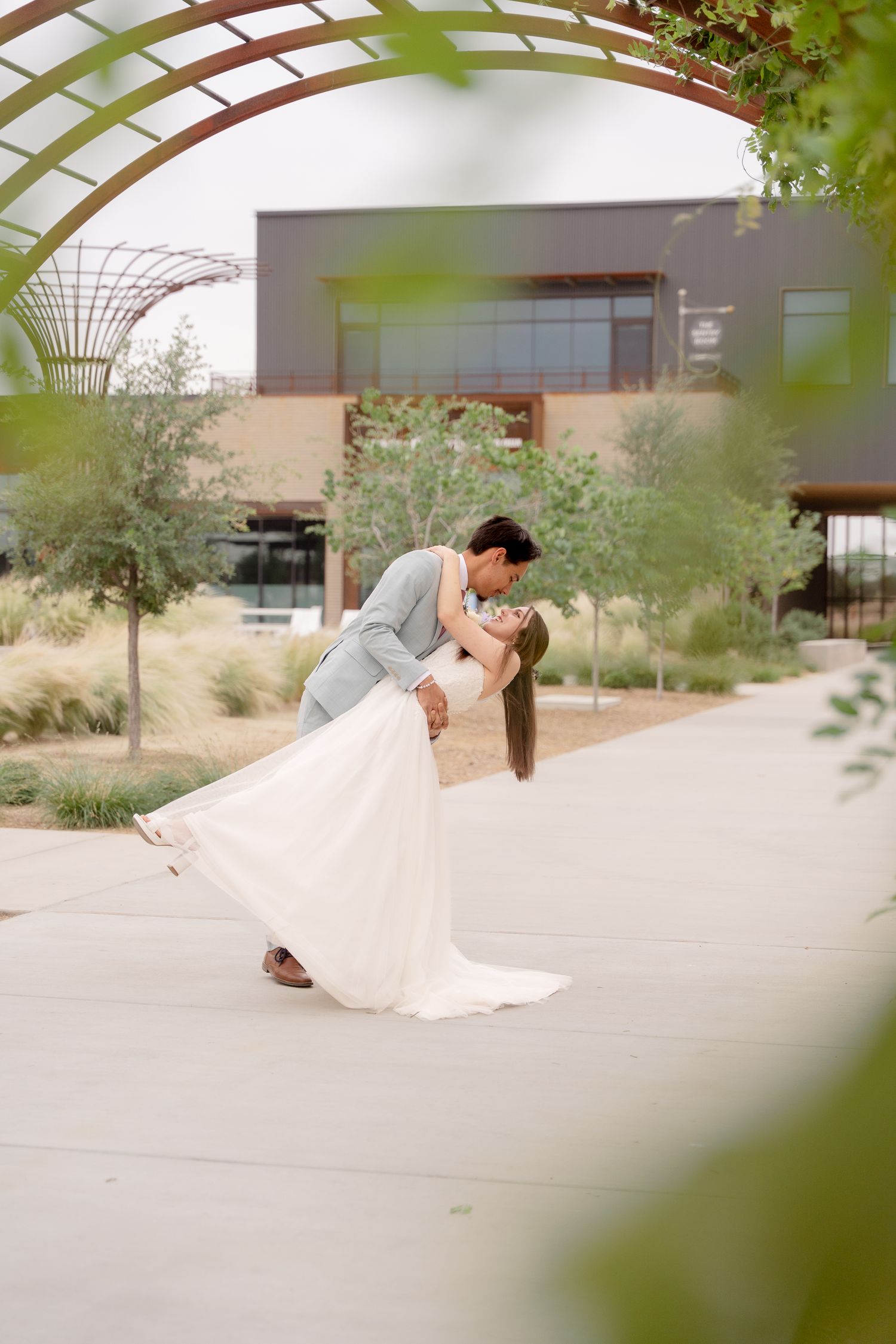 A couple shares an intimate dip kiss under a rustic wooden pergola surrounded by desert landscaping.