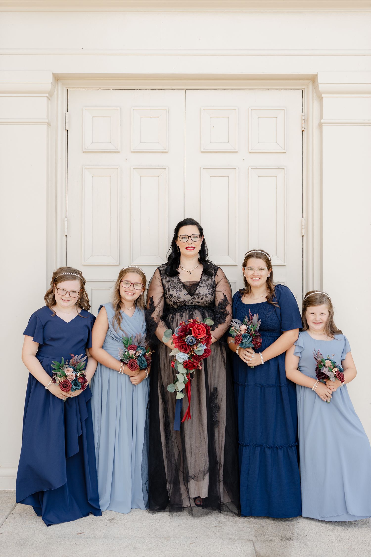 A bridal party wearing navy and light blue dresses pose together against a white wall.