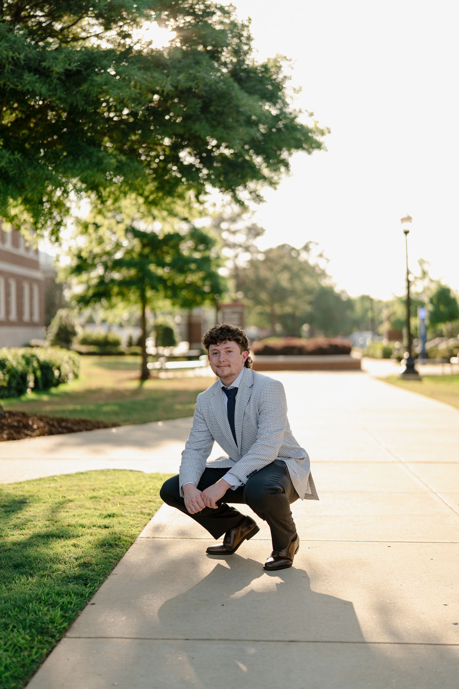 Smiling young man in gray blazer crouches on a sunlit campus walkway surrounded by green trees.
