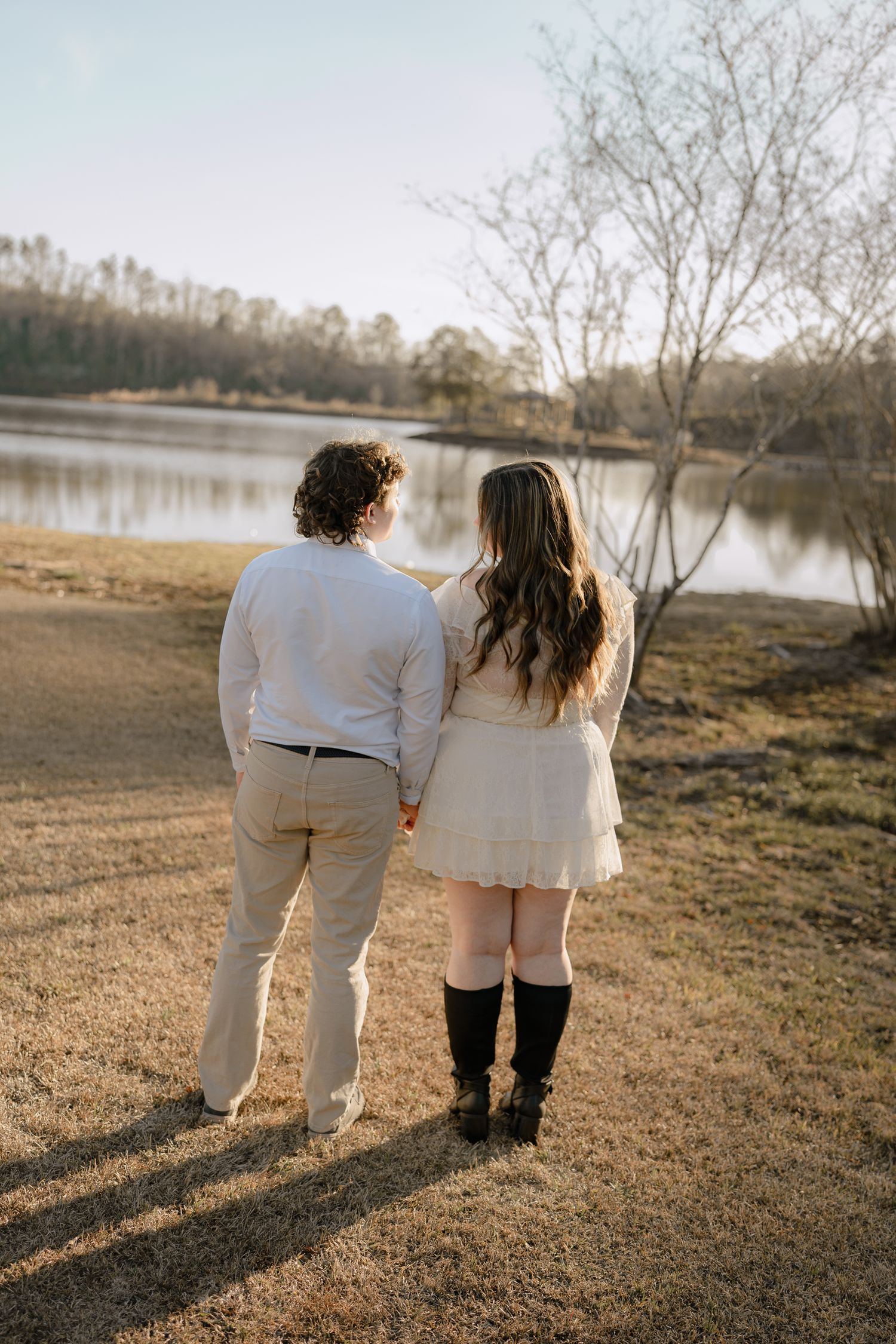 Couple holding hands walking toward a peaceful lake at sunset, woman in white dress and black knee-high boots.