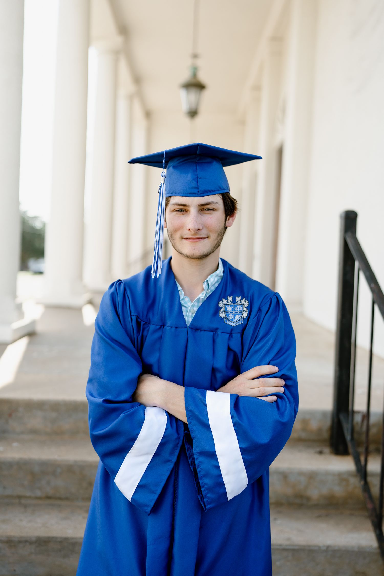 Graduate in blue cap and gown smiles confidently while standing in columned walkway.