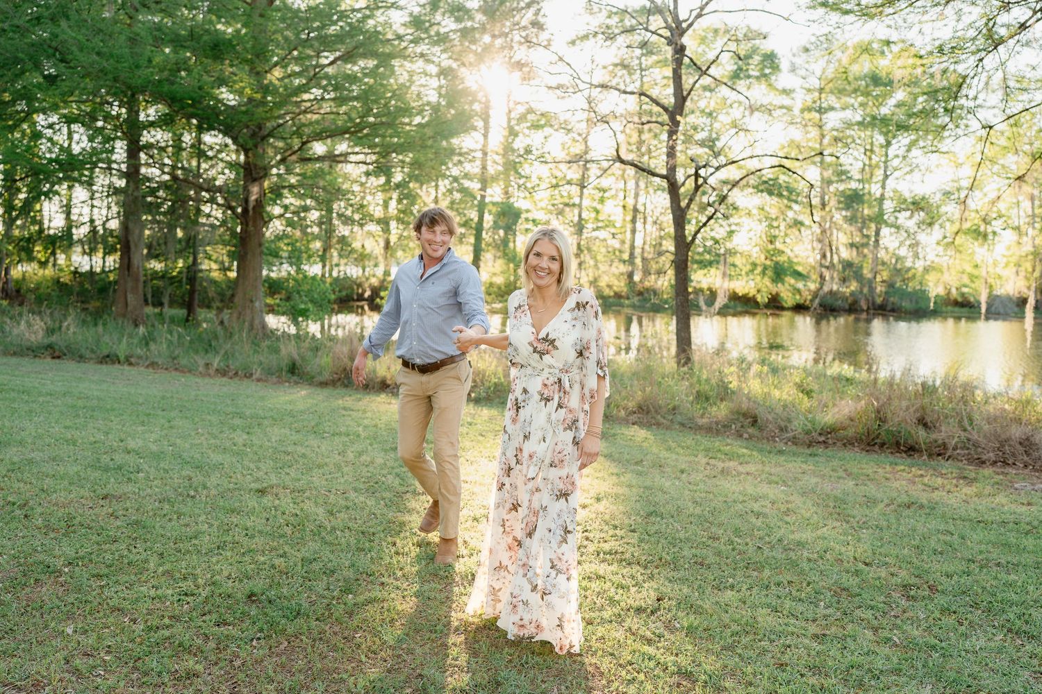 A couple walks together on grass at sunset with trees and a lake in the background.