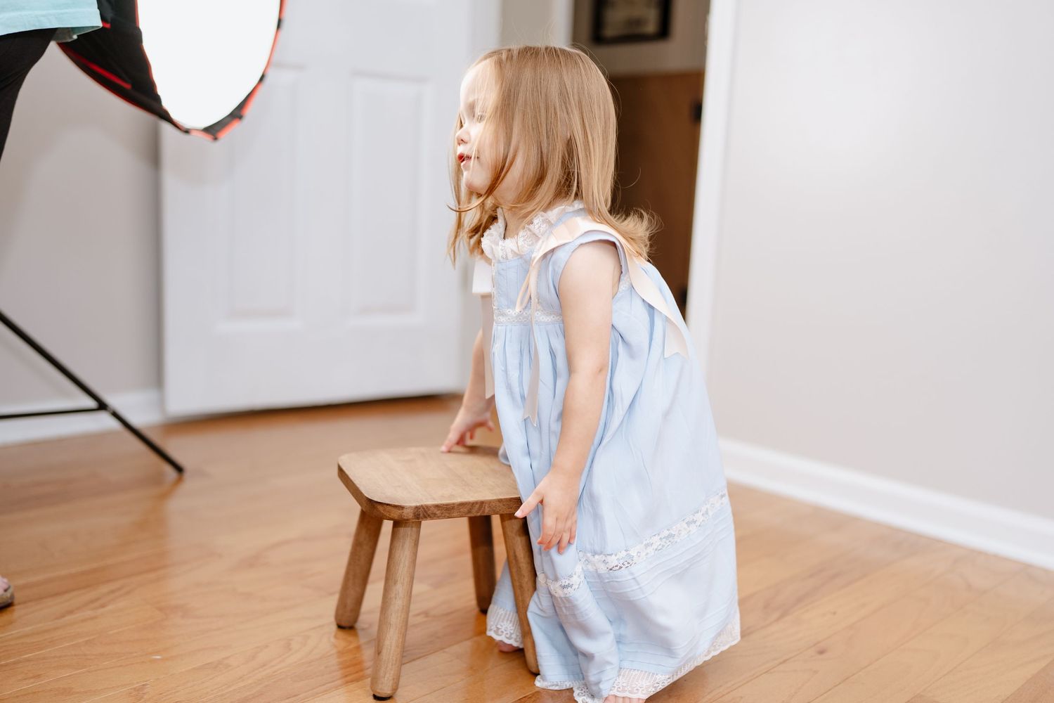 Child in blue dress standing next to wooden stool during photo session.