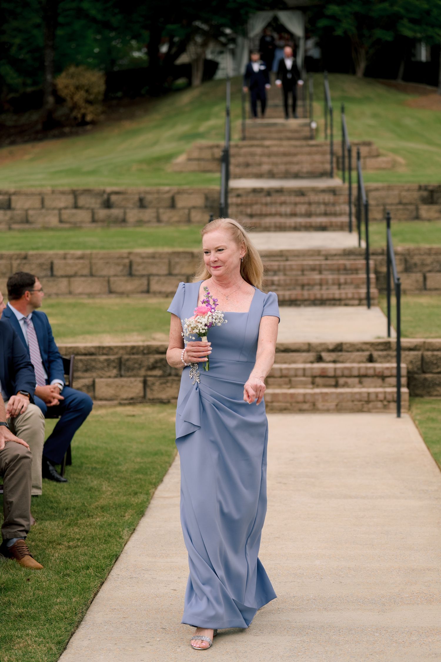 Bridesmaid in blue gown walks down outdoor aisle carrying wildflower bouquet at garden wedding.