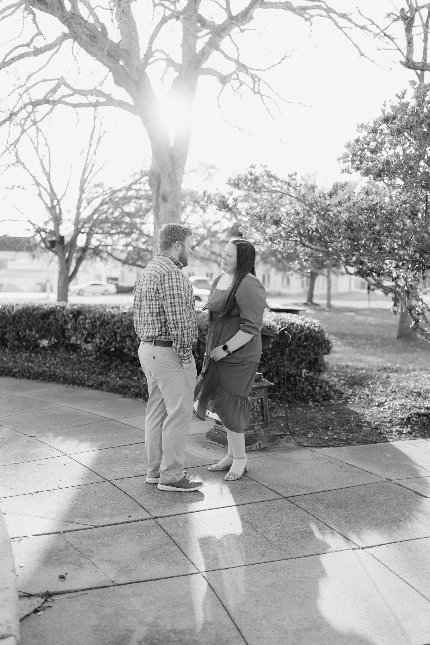 Black and white photo of couple holding hands on sidewalk, surrounded by trees in soft backlit sunlight.