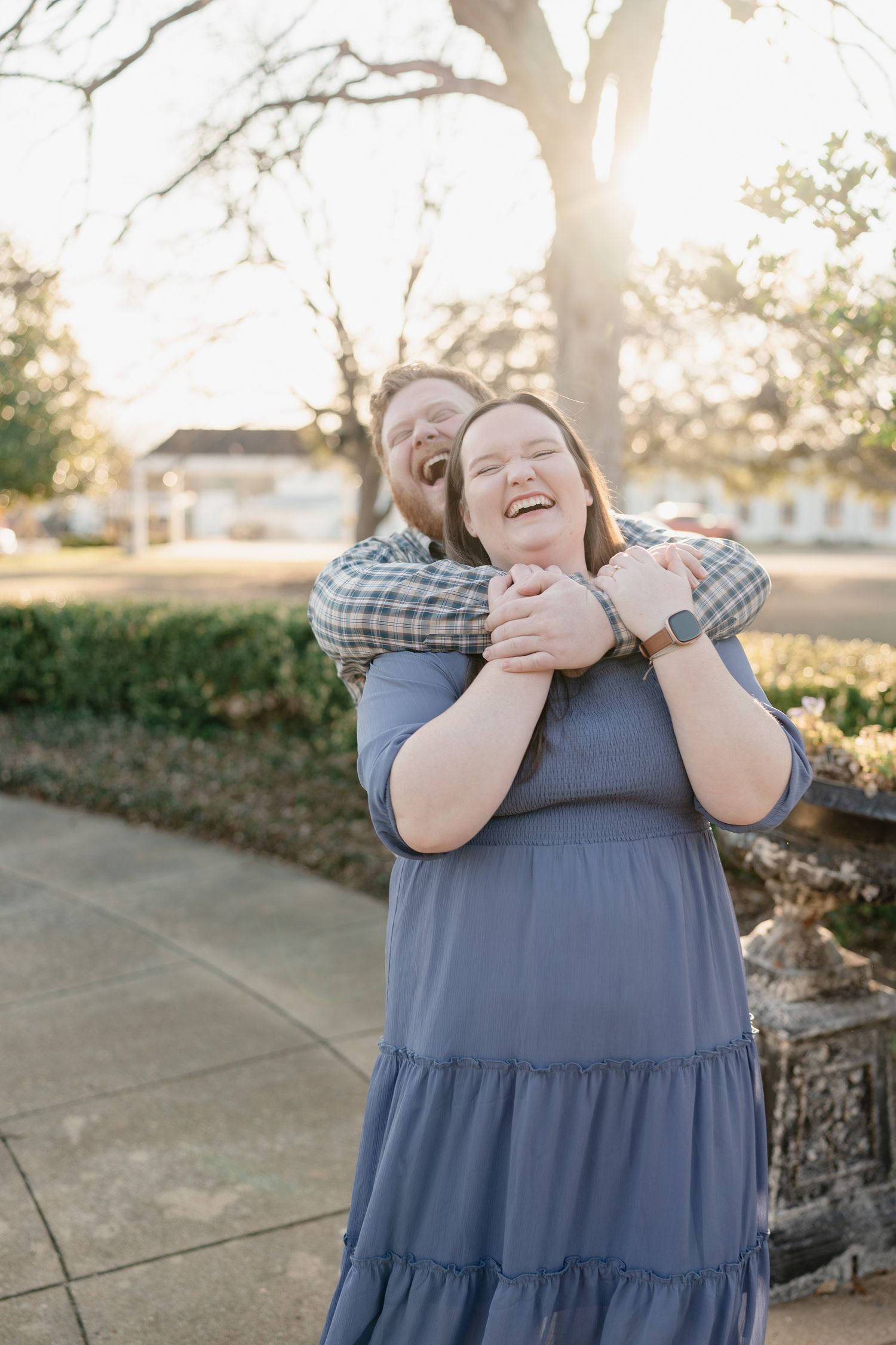 A couple laughs joyfully outdoors at golden hour, man hugging woman in blue dress from behind on a sunlit pathway.