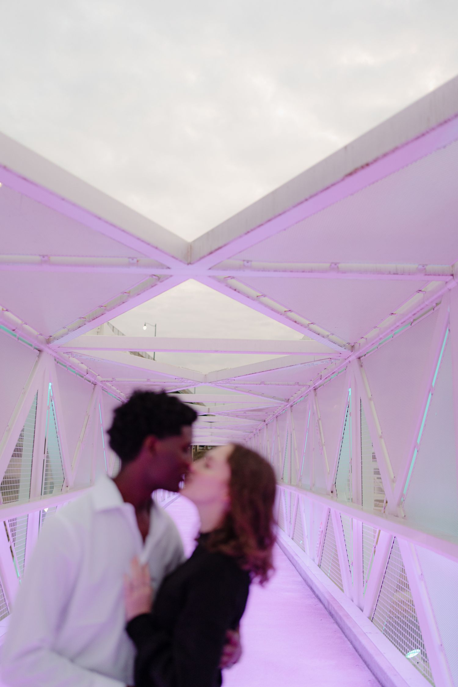 Couple sharing a kiss on a purple-lit pedestrian bridge at dusk.