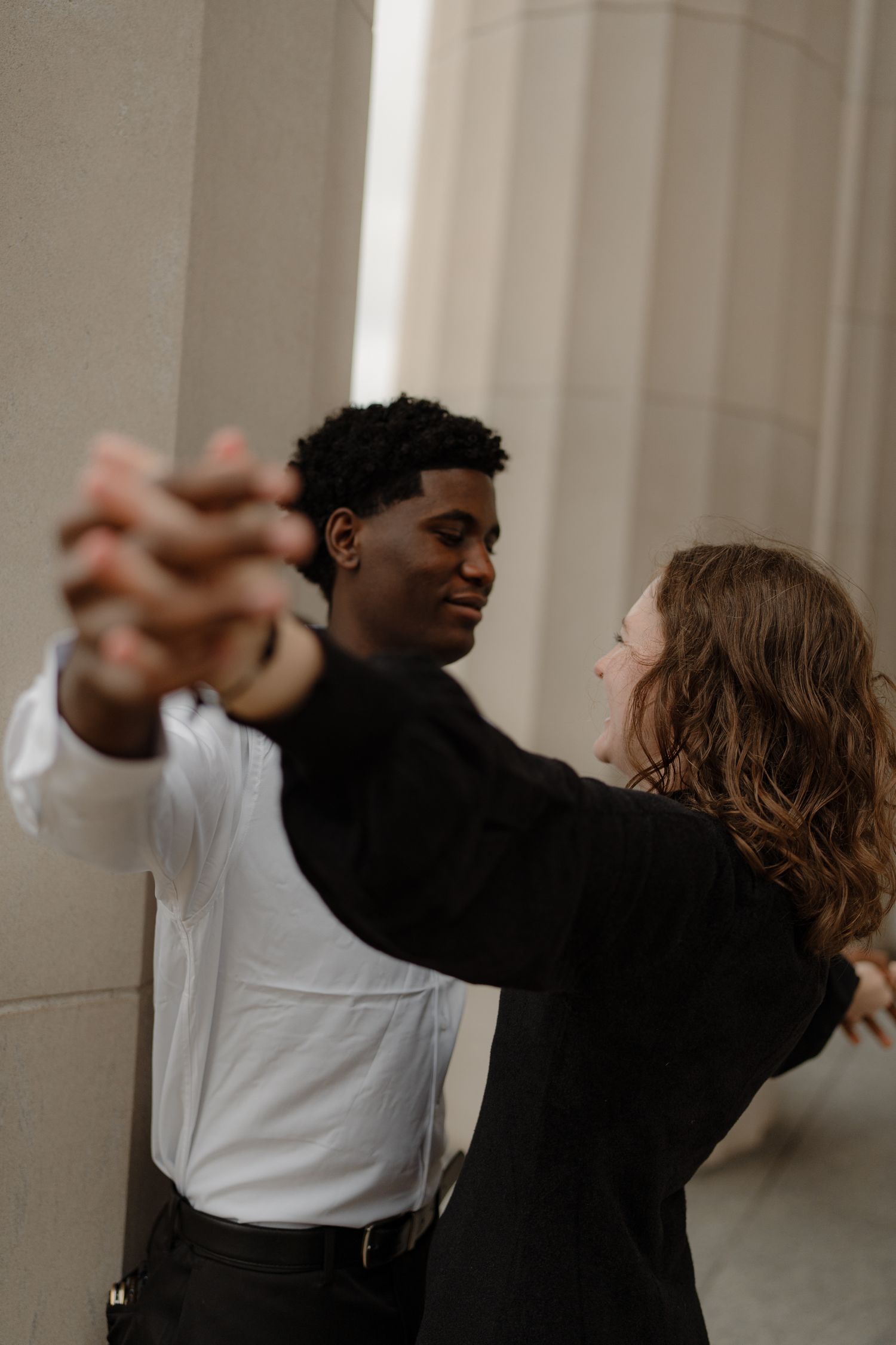 Young interracial couple holding hands and dancing near large stone pillars outdoors.