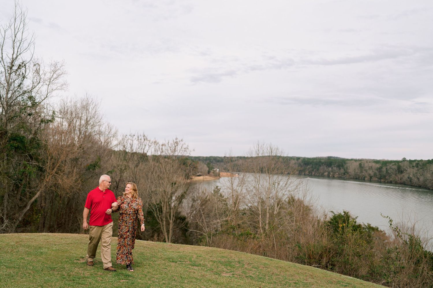 Couple in red and floral attire walking hand in hand along a grassy hill by a river.