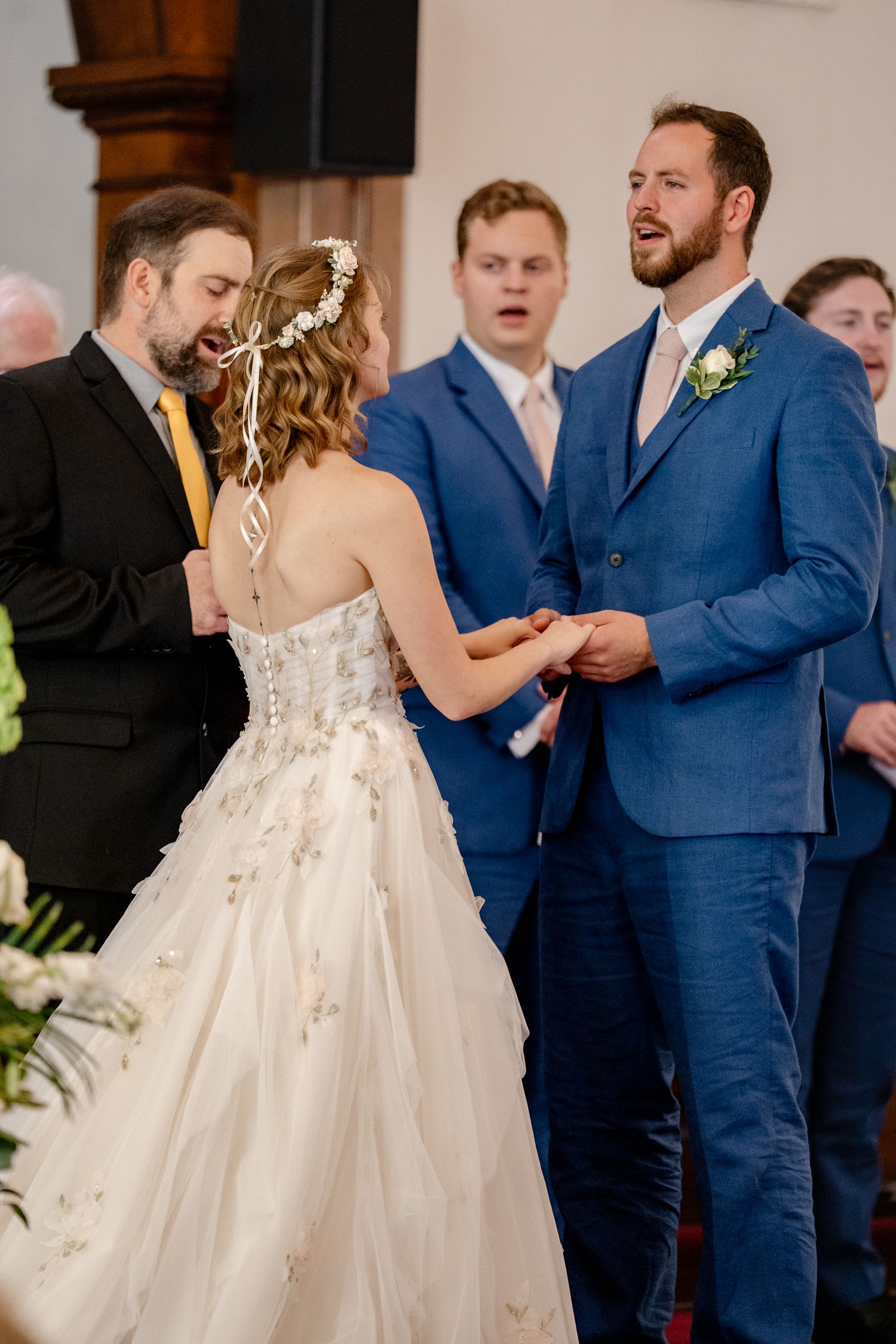 Wedding ceremony scene with bridesmaids in navy blue dresses and groomsmen in matching suits during ring exchange.