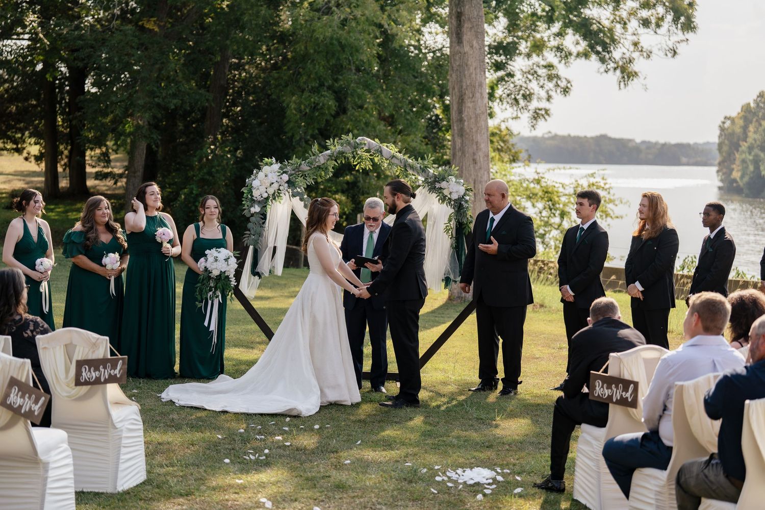 Wedding party gathers outdoors near a lake for a scenic ceremony under tall trees at sunset.