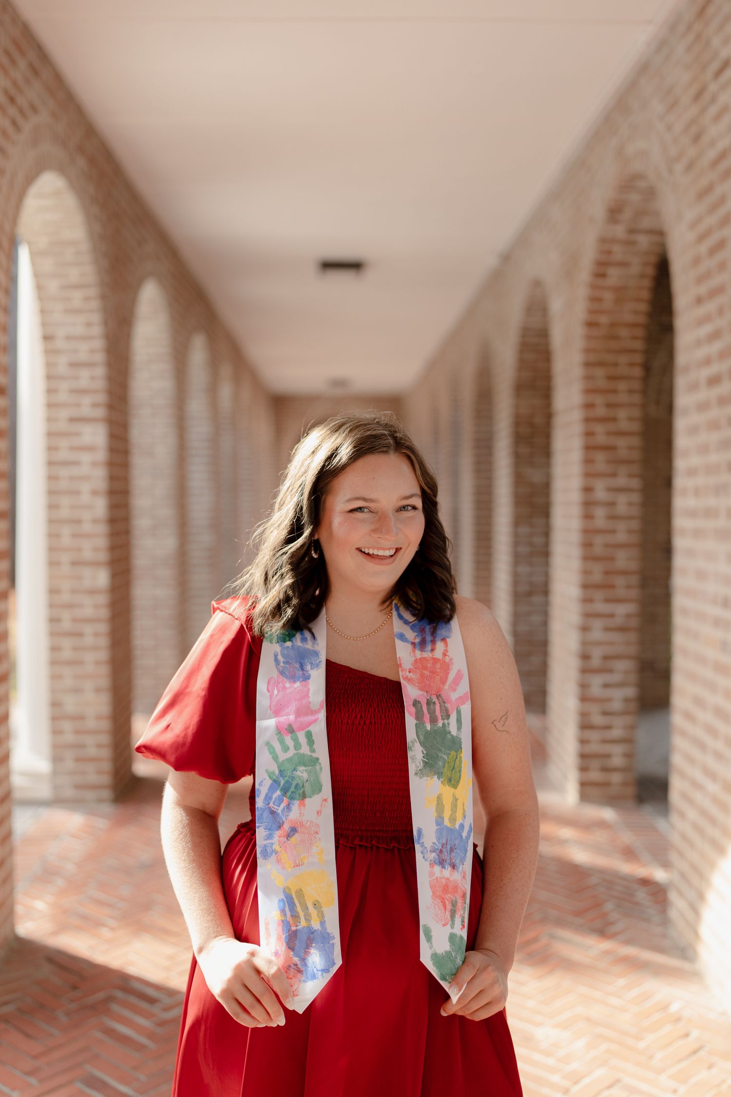 A person in a red dress and colorful vest stands in a brick-lined hallway with arches and natural lighting.