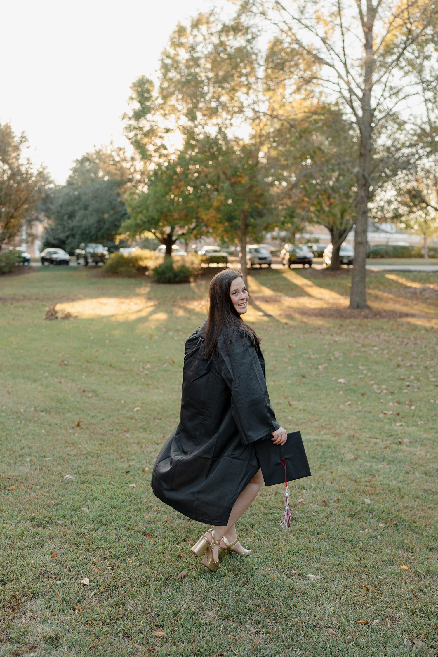 A person in a black graduation gown spins around on a grassy lawn during sunset, creating a flowing motion effect.