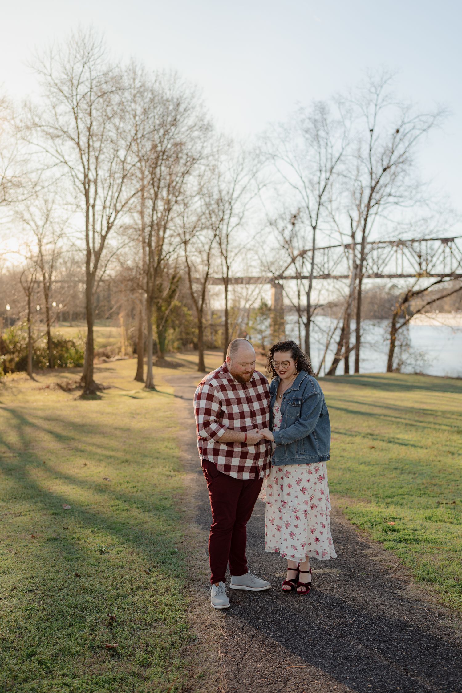 A couple walks together on a path through a scenic park with bare trees and a lake in the background.