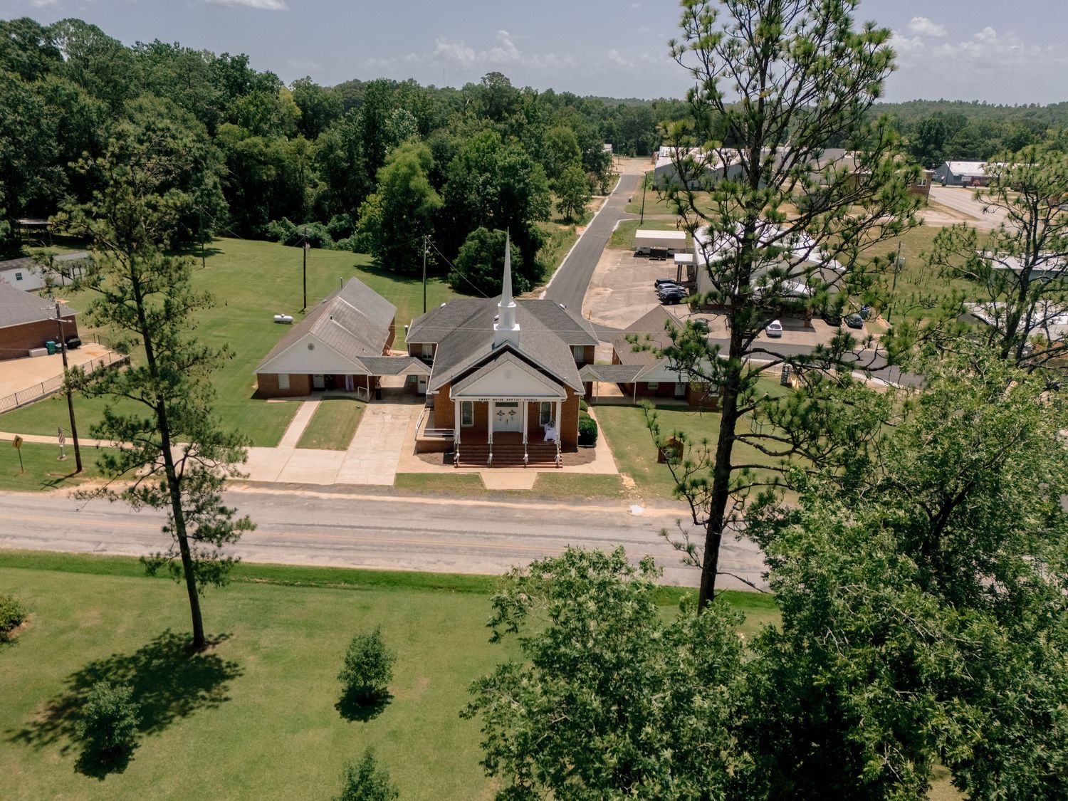 Aerial view of small town church with steeple surrounded by green trees on sunny day.