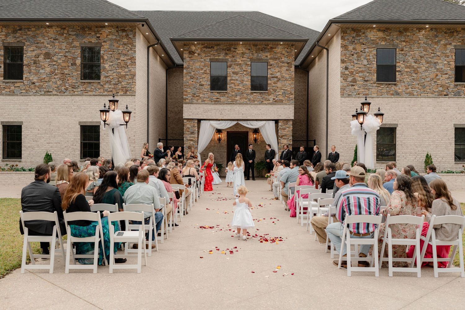 Outdoor wedding ceremony with white chairs and rose petals lining the aisle at a stone building venue.