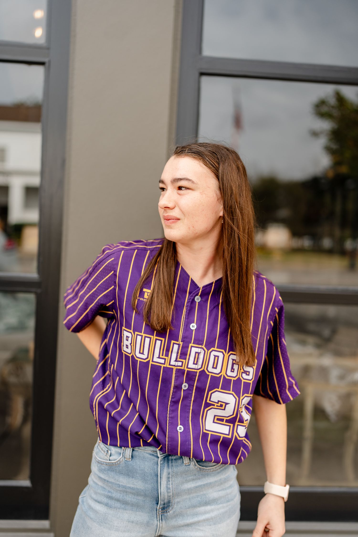 Person in purple striped baseball jersey and denim poses against grey building exterior.