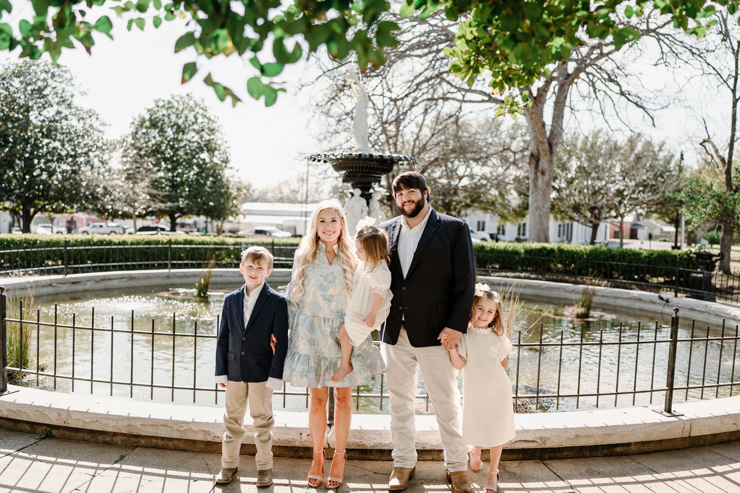 Family group standing along outdoor walkway near fountain in coordinated casual outfits.