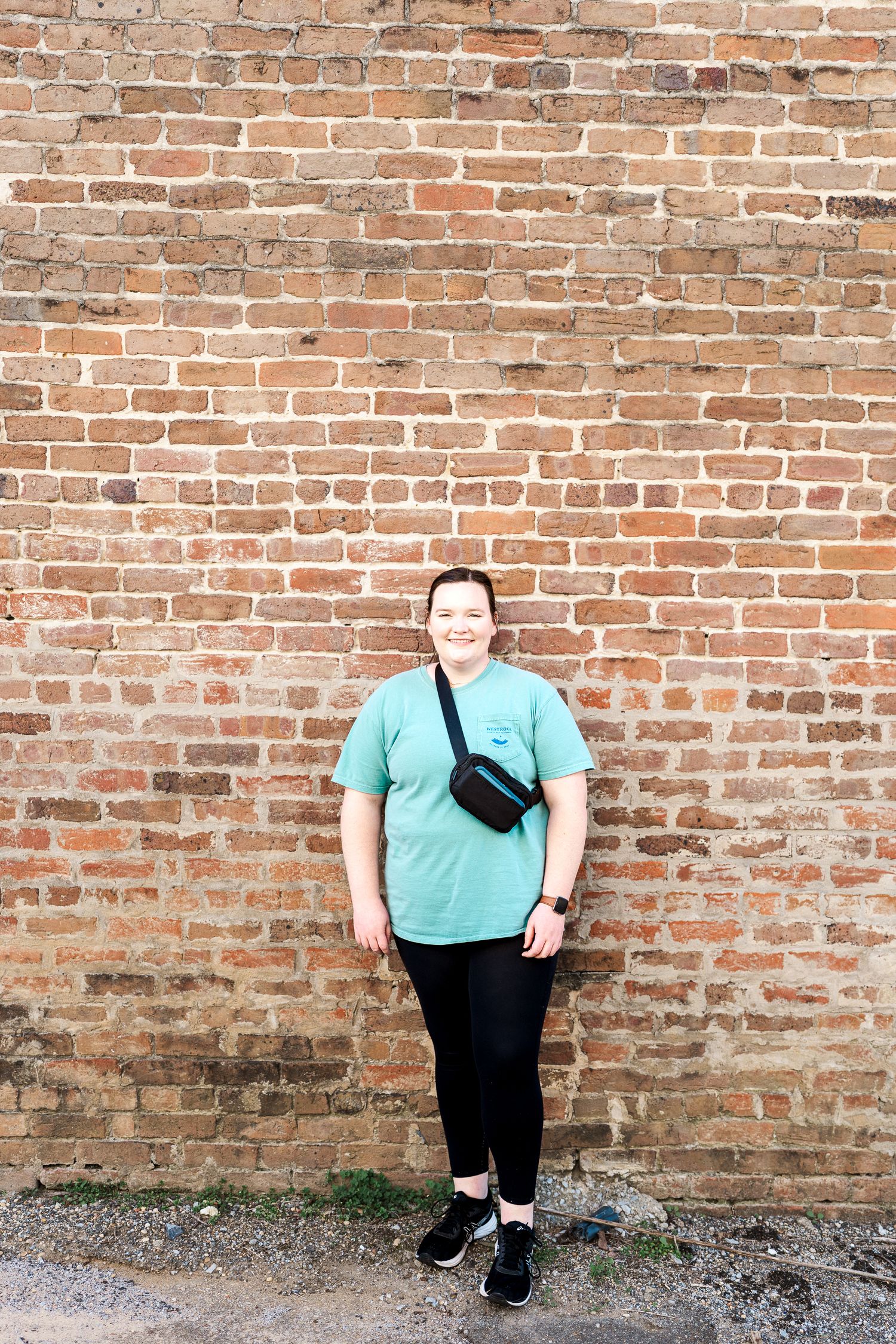 Person in teal shirt and black pants poses against weathered brick wall with fanny pack.