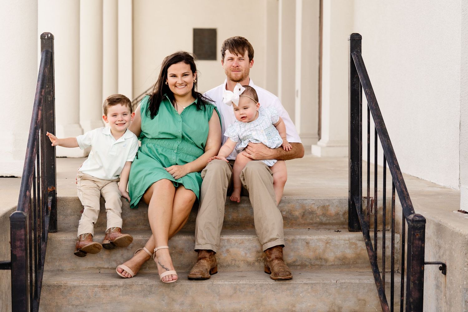 Family of four sitting together on outdoor steps between classic white columns.