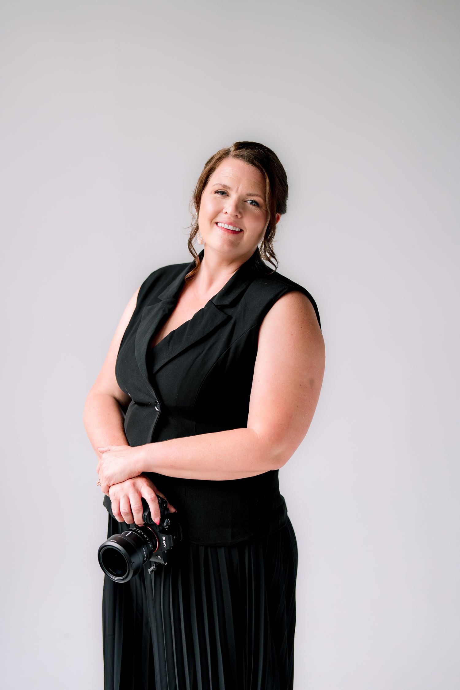Woman in black sleeveless dress holding a professional camera, smiling confidently against a light gray background.
