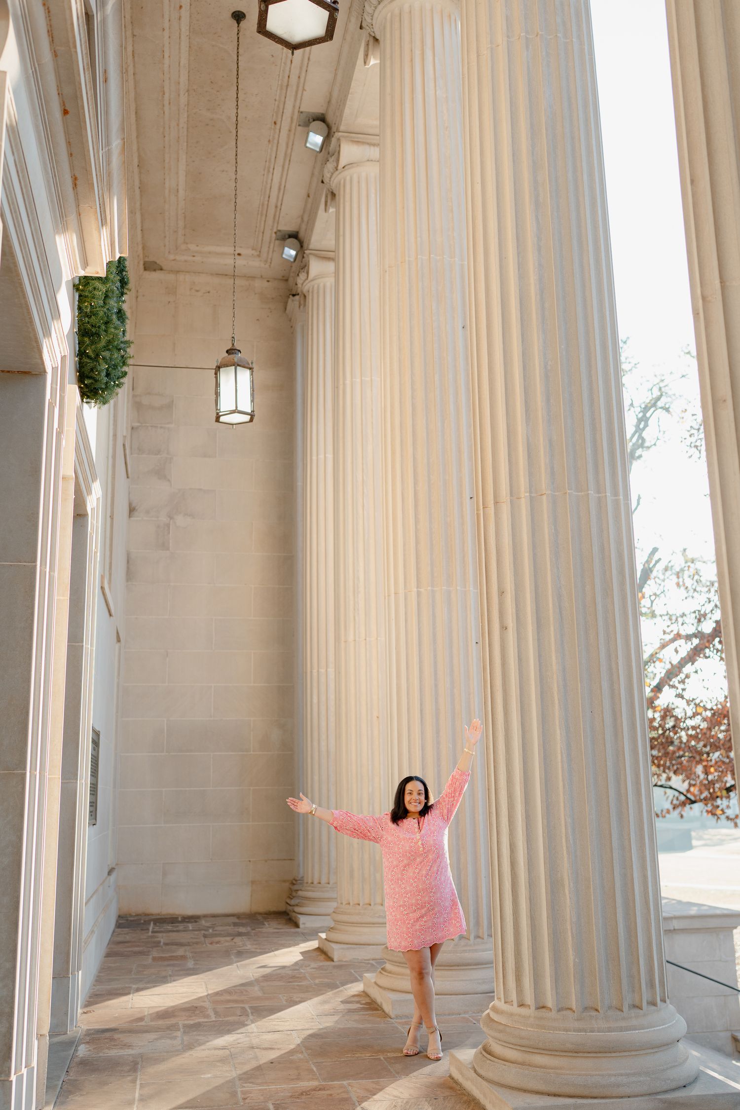 Graduate in pink dress celebrates between towering white columns with natural lighting.