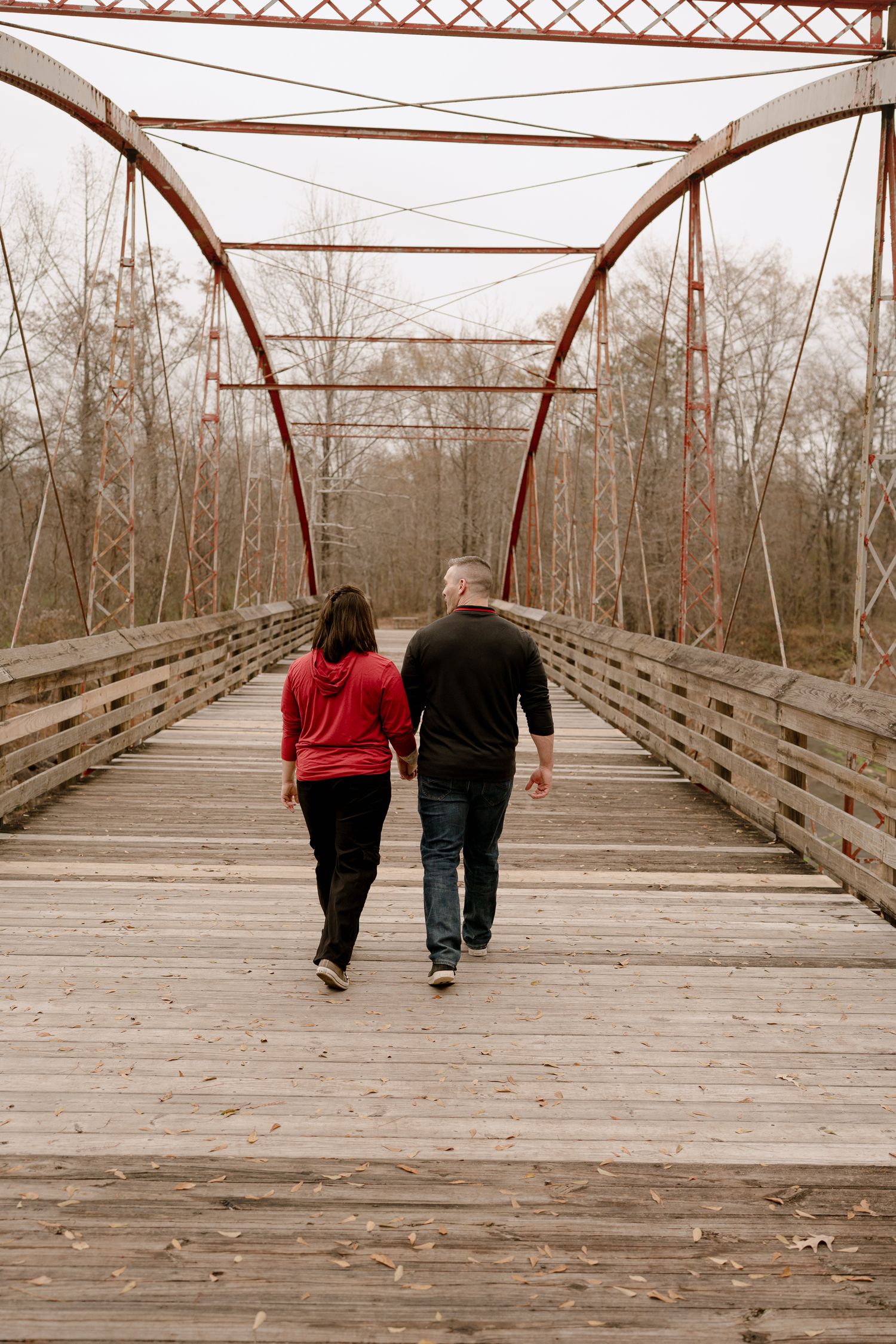 A couple walks hand in hand across a rustic wooden bridge with metal trusses on a cloudy day.