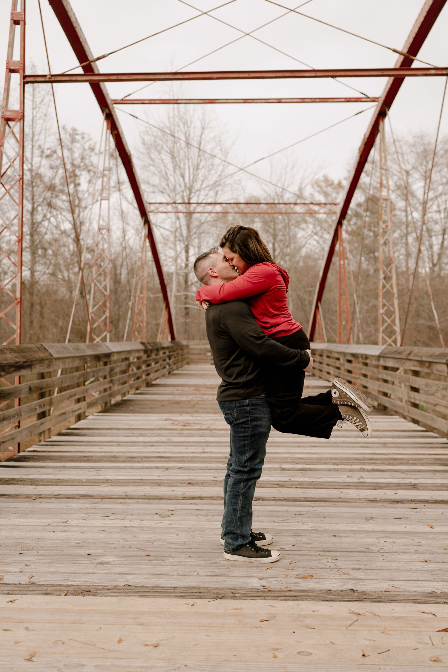 A couple shares a playful embrace while standing on a wooden bridge with metal beams overhead.