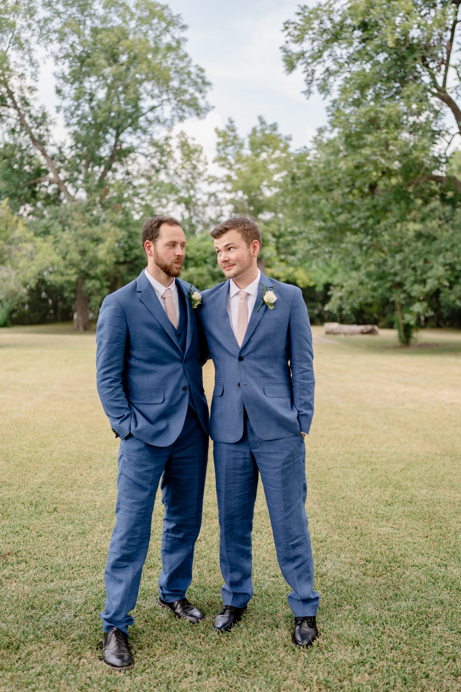Two people in matching navy blue suits stand together on a grassy lawn with trees in the background.