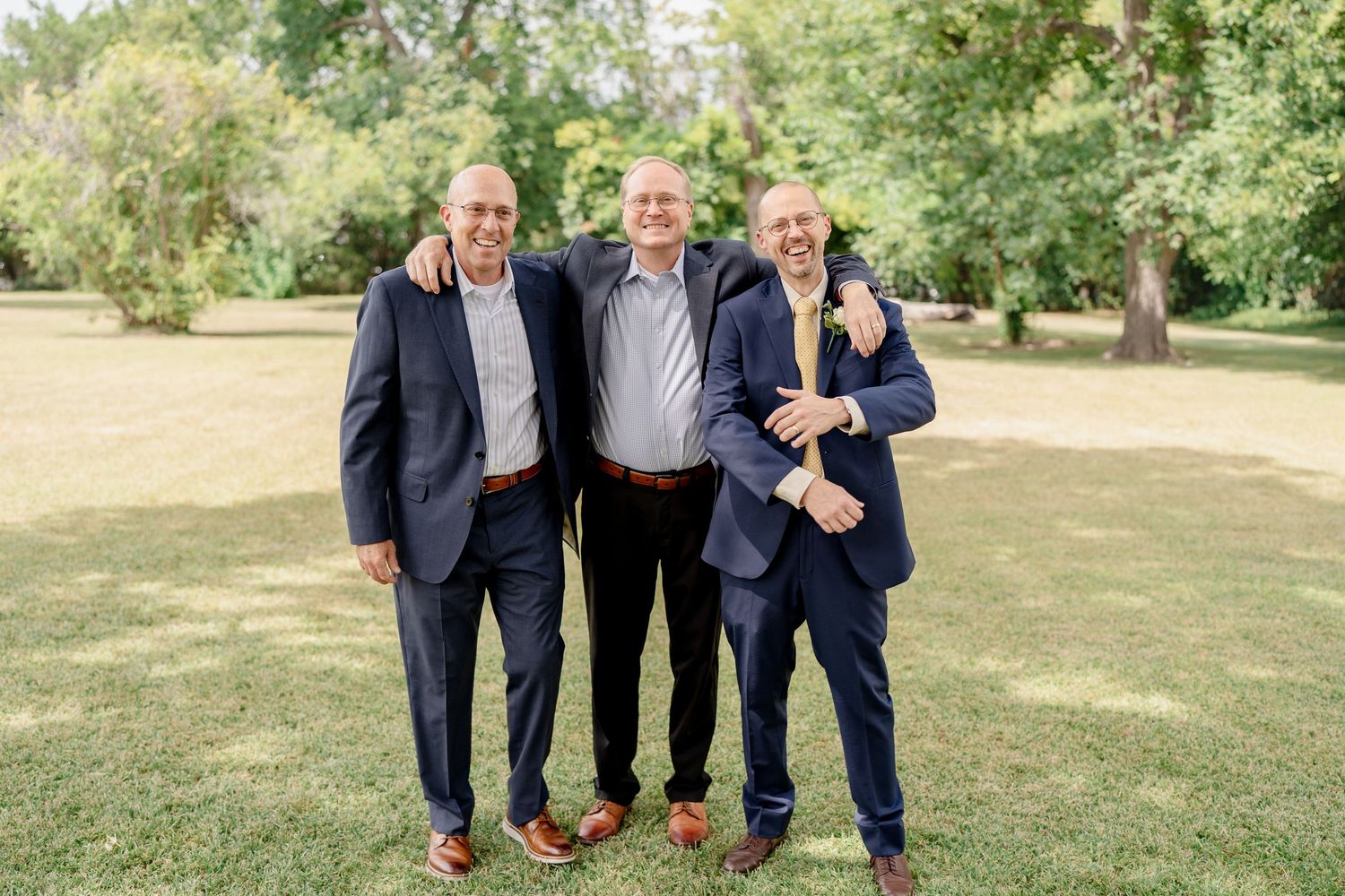 A group in navy suits poses together on a grassy lawn during a sunny outdoor gathering.