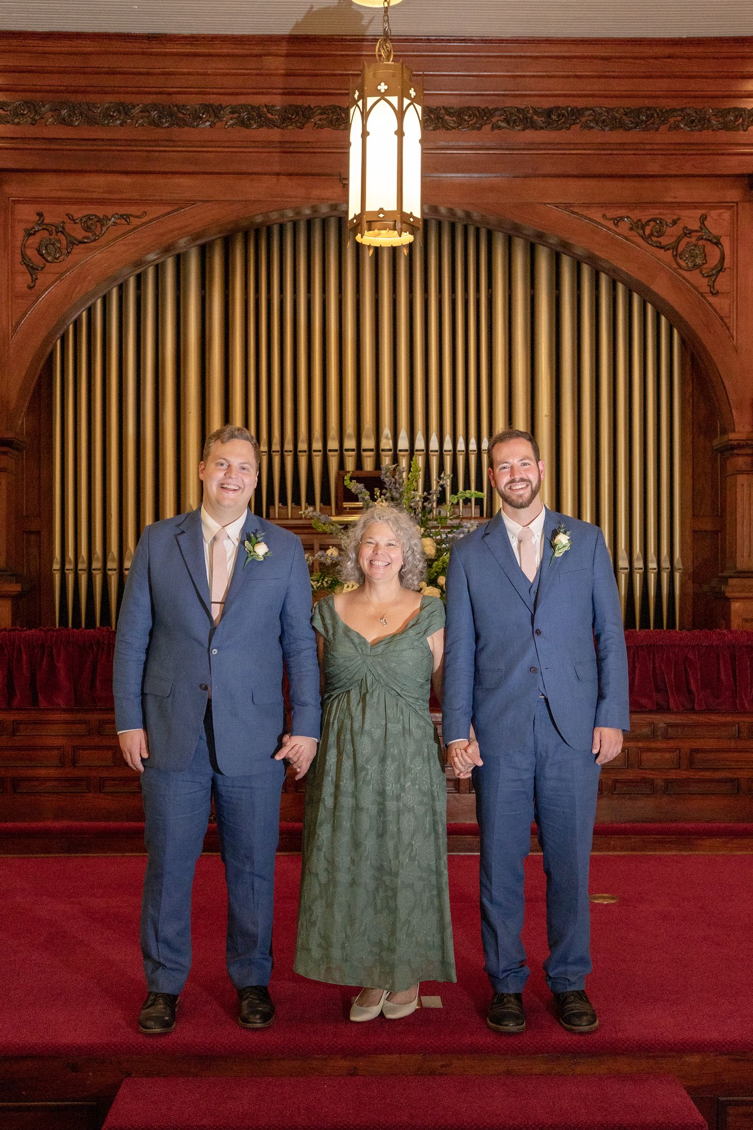 Three people stand together in formal attire against an ornate church backdrop with organ pipes and red carpet.