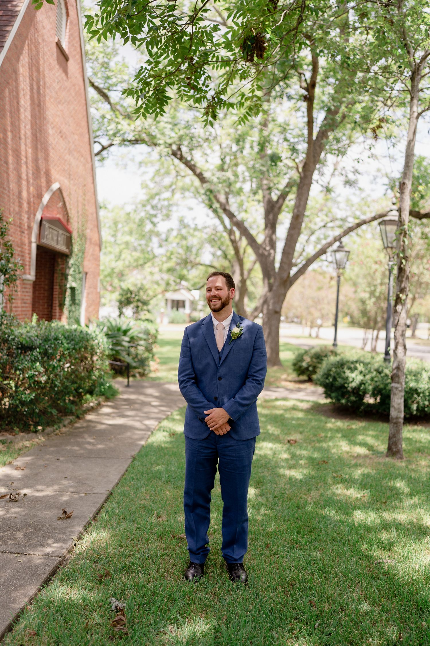 A person in a navy blue suit stands on a path near a brick church surrounded by green trees and landscaping.