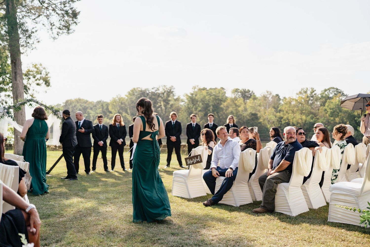 Wedding party members stand in line outdoors during sunset for group photos in a natural setting.
