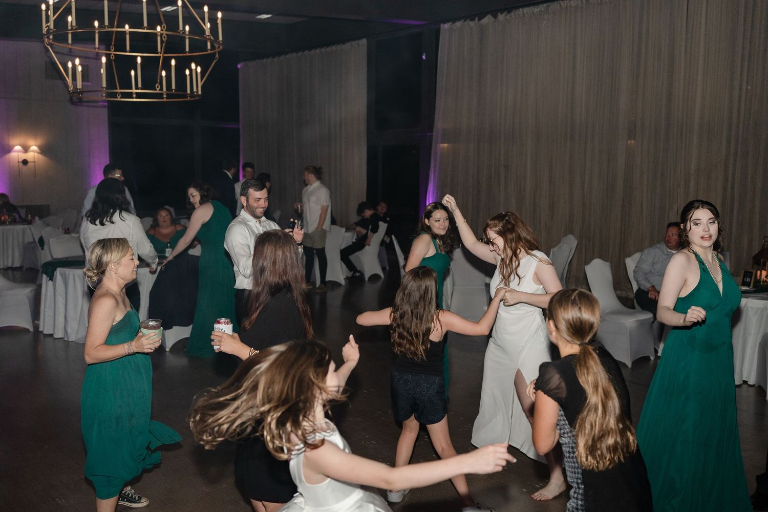 Wedding guests dancing and celebrating on a dimly lit dance floor with emerald green bridesmaids dresses.