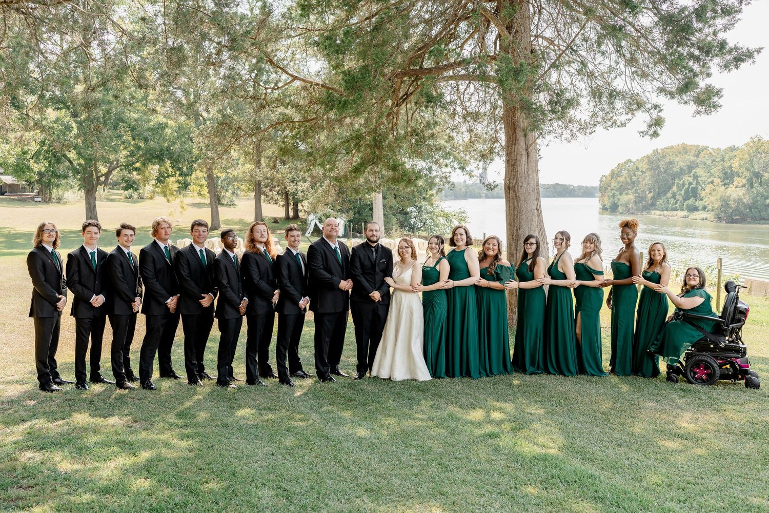 Wedding party members in black and emerald green attire pose outdoors under trees near a waterfront setting.
