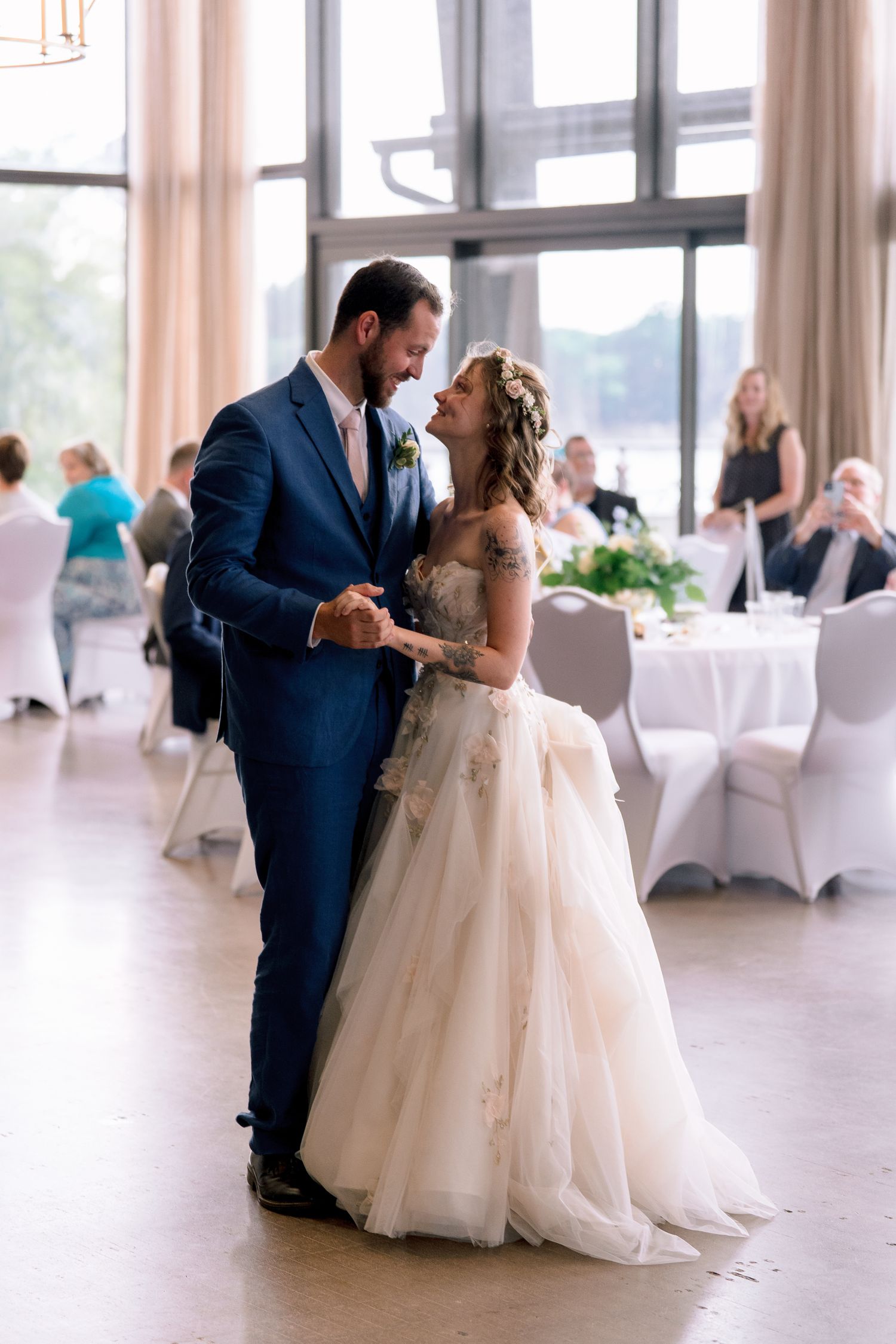Bride and groom share their first dance in an elegant wedding reception hall with large windows and white-draped tables.
