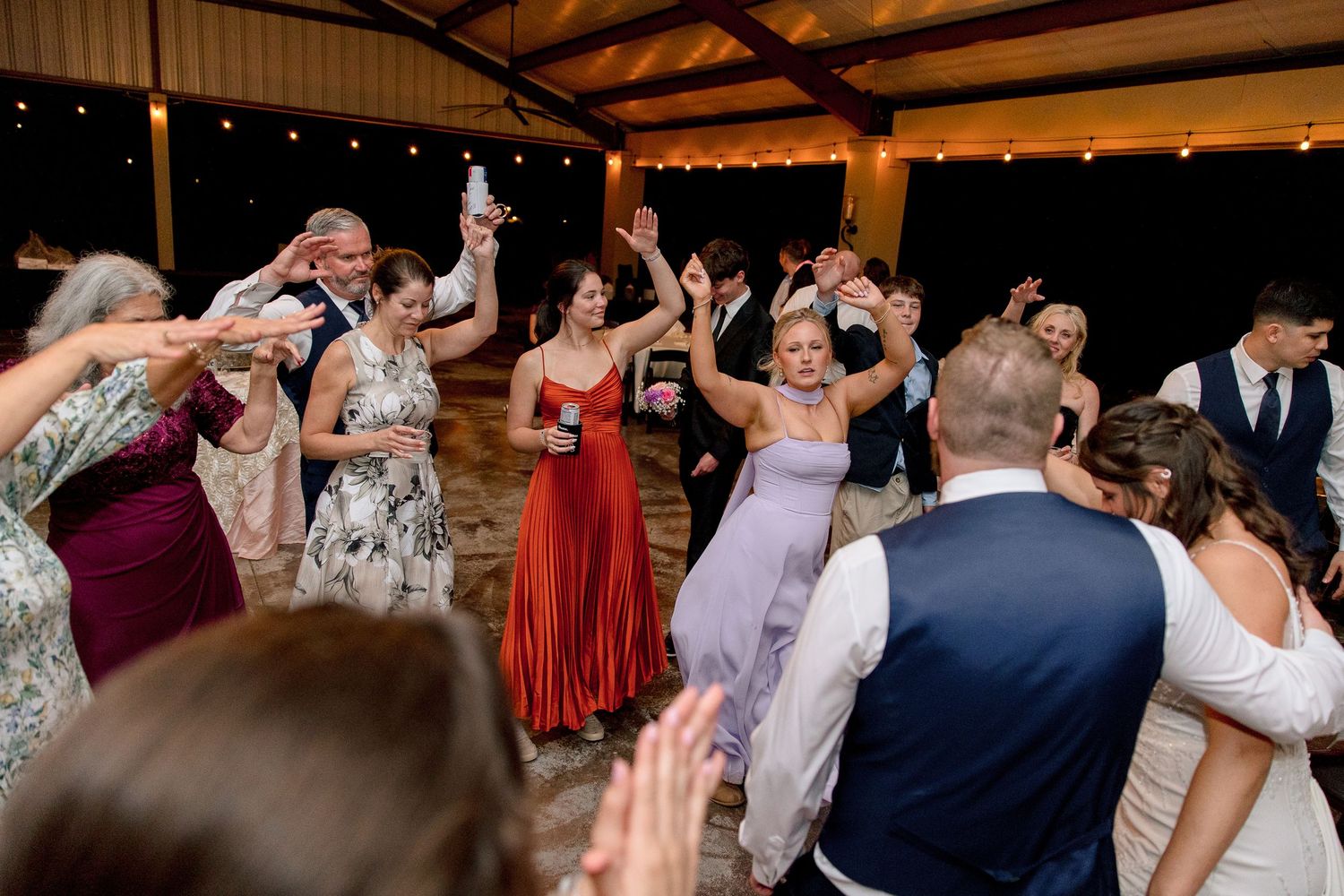 Wedding guests fill the dance floor with hands raised during lively indoor reception celebration under string lights.