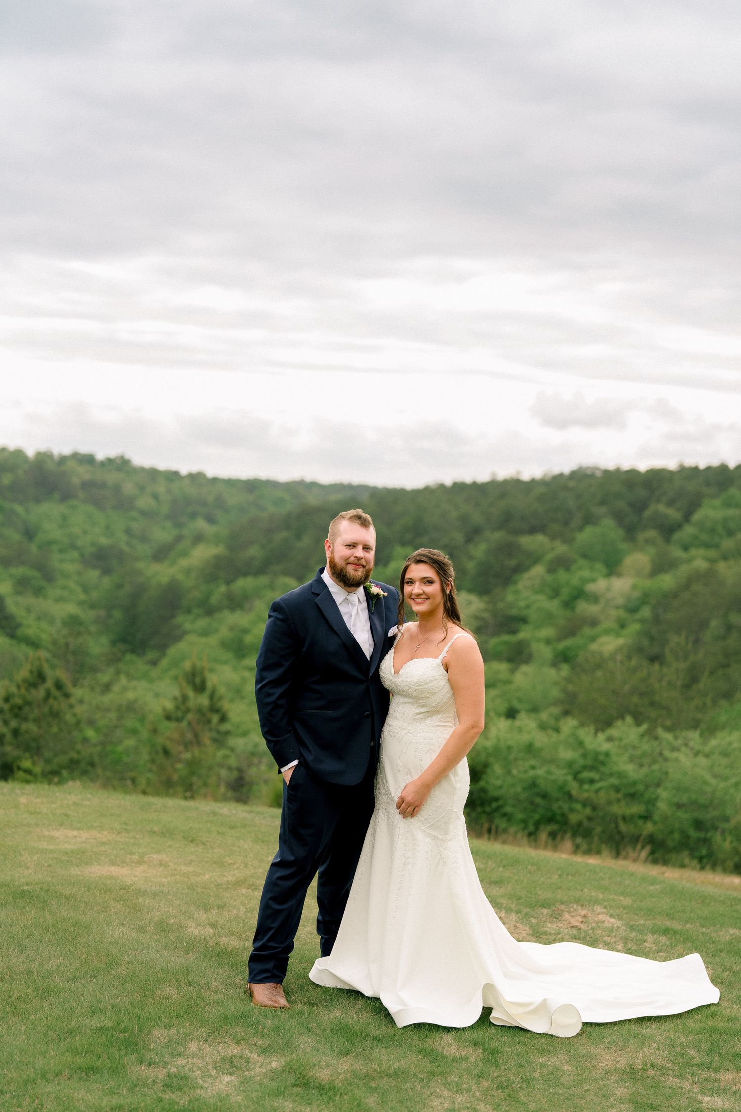 Bride and groom pose together on a lush green hillside with forest backdrop on their wedding day.