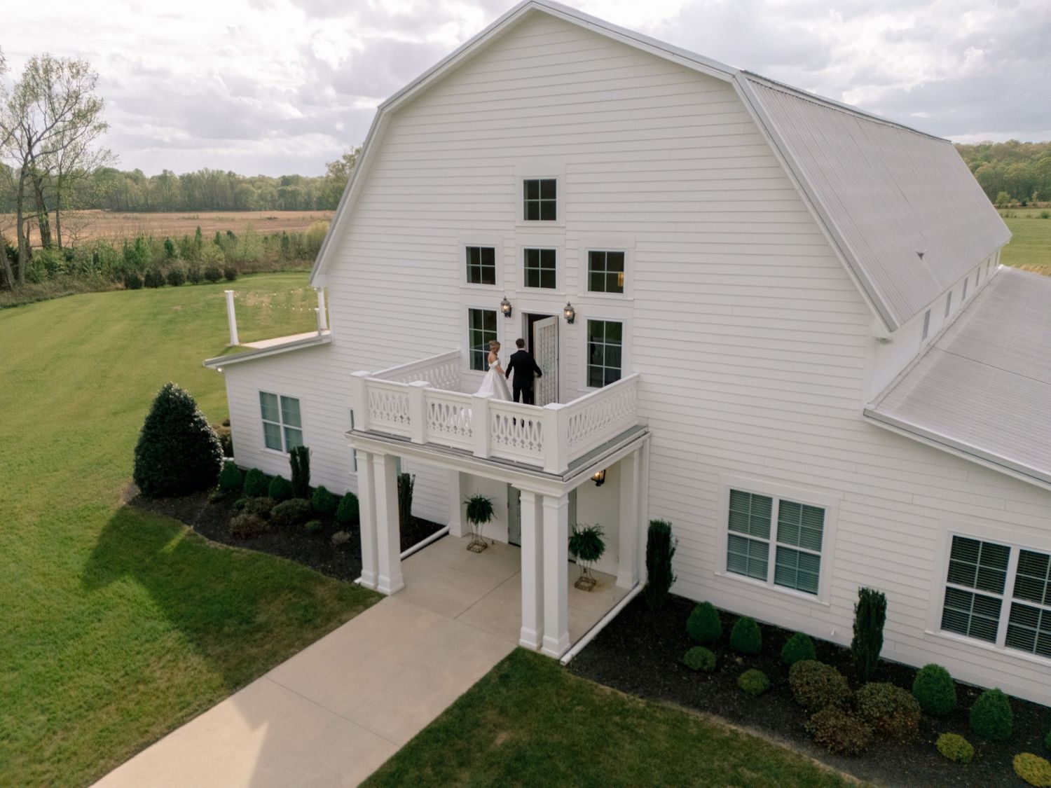 Aerial view of a white barn wedding venue with a couple on the balcony, surrounded by lush greenery and open fields.