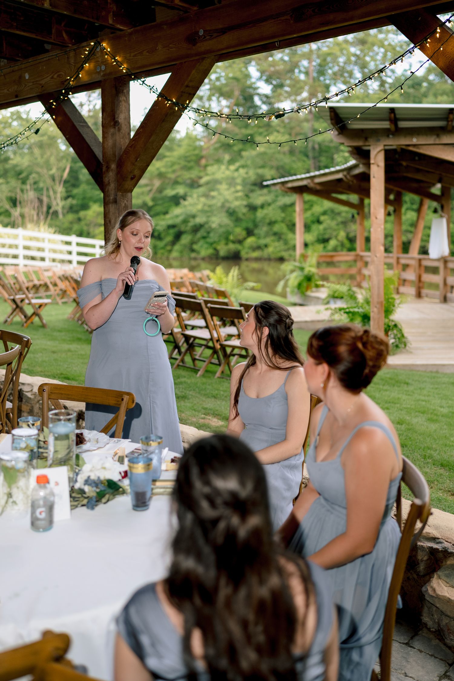 Bridesmaid gives speech at outdoor wedding reception under rustic pavilion while others in blue dresses listen.