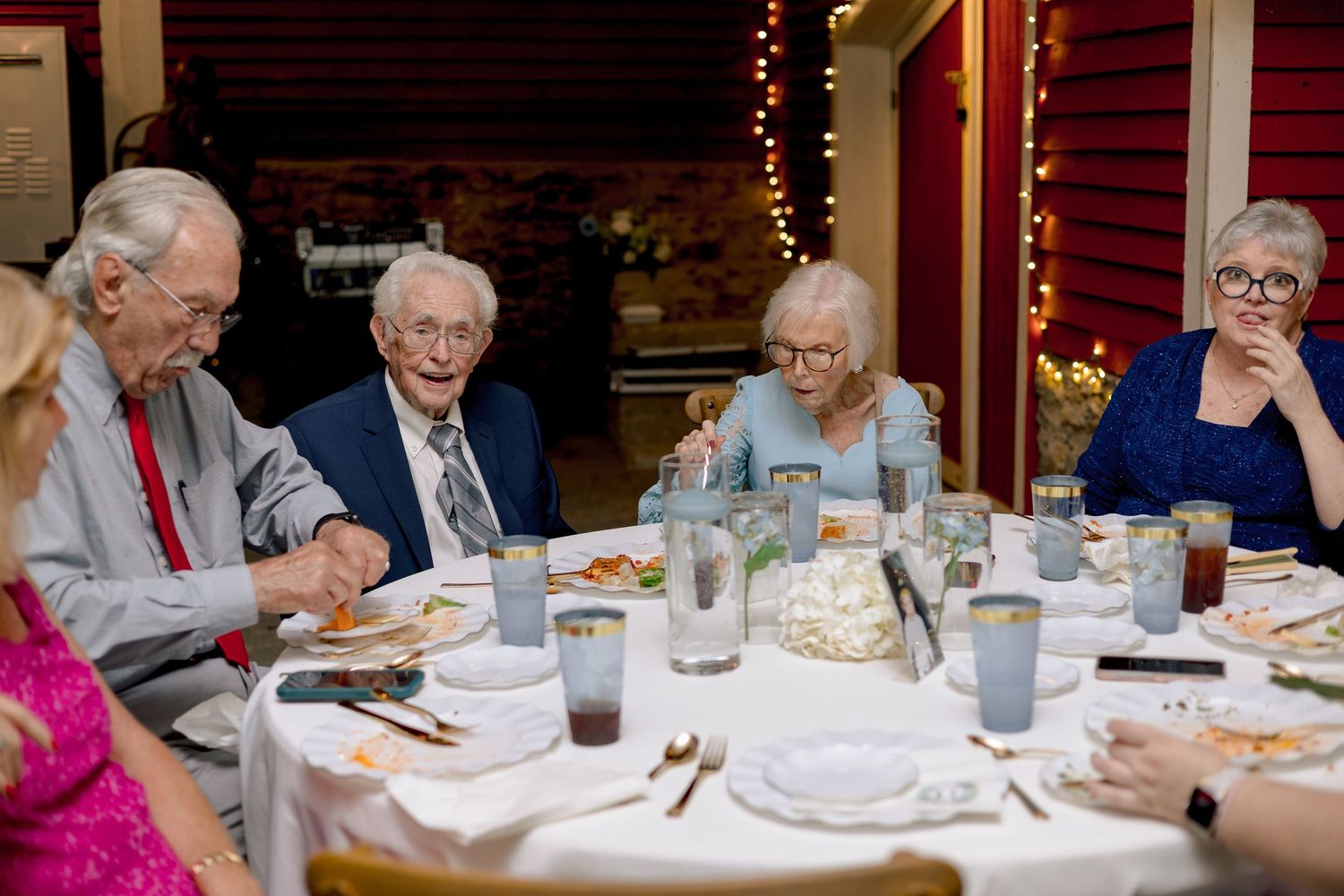 Elderly wedding guests enjoying dinner at round table with string lights at rustic reception venue.