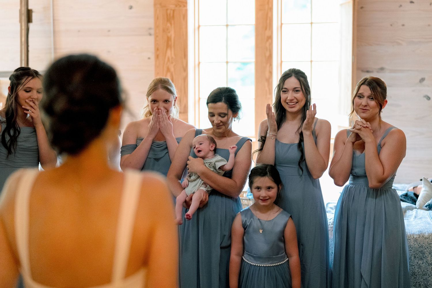 Bridesmaids in blue-gray dresses react emotionally as bride reveals her wedding look inside a rustic barn venue.