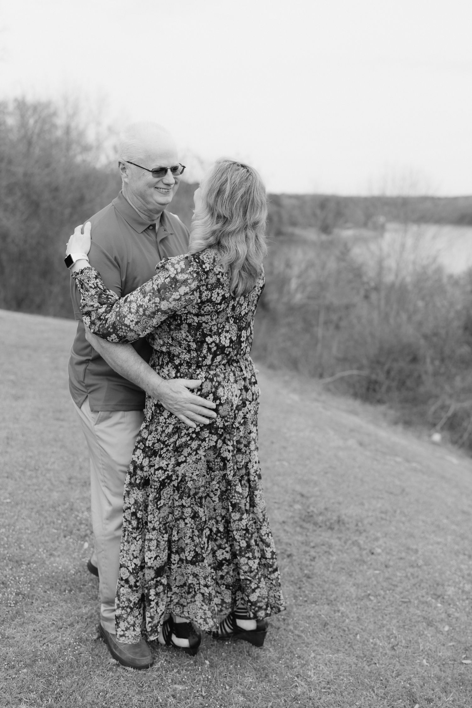 Older couple embraces and dances together outdoors near a lake in black and white.