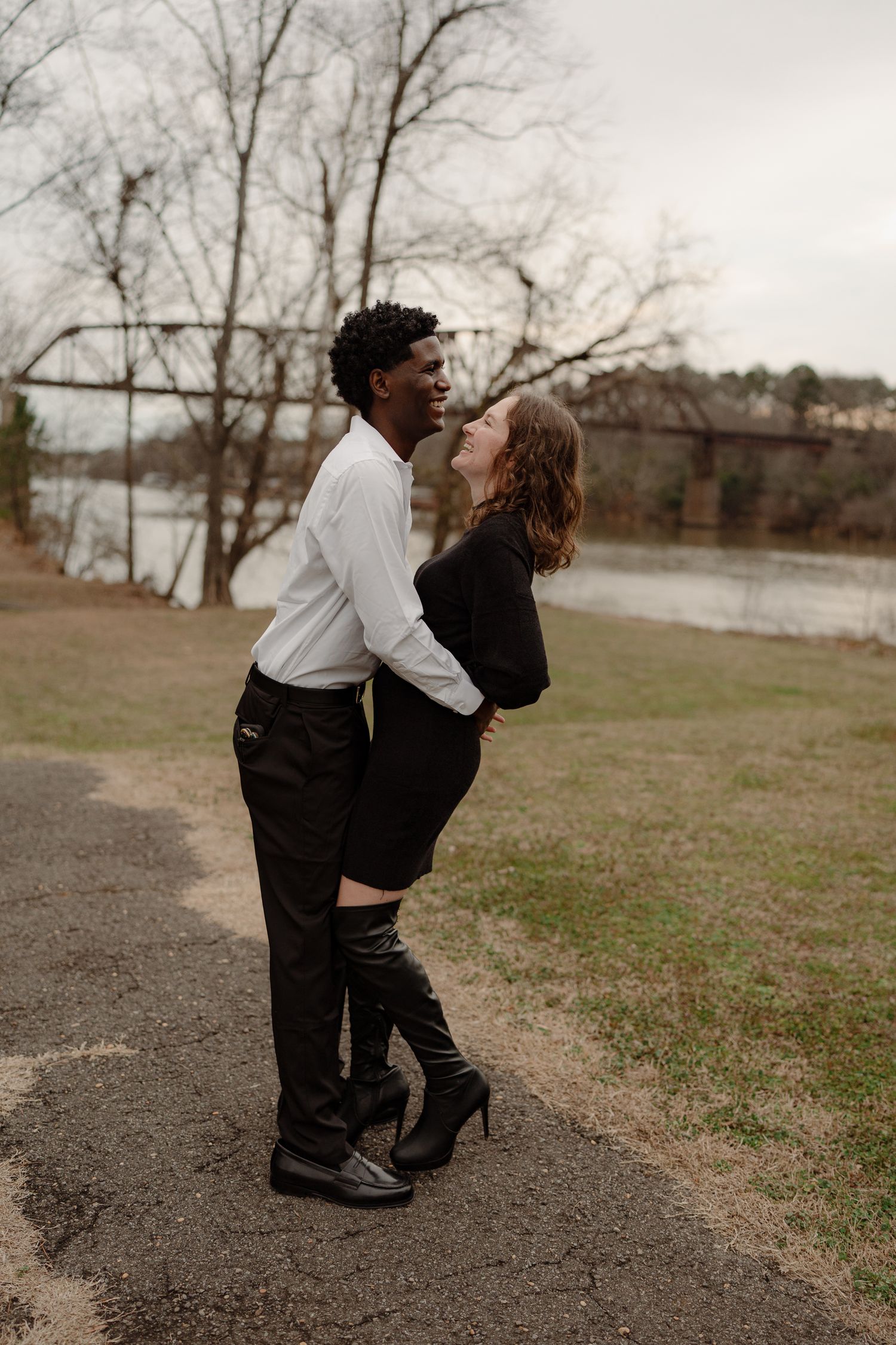 An interracial couple shares a romantic moment, gazing at each other near a riverside bridge.