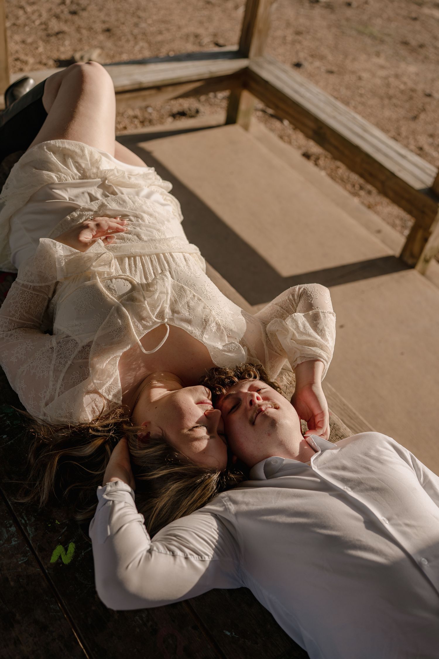 Couple lying together on a wooden swing, woman in white lace dress and man in white shirt, sharing a tender moment.