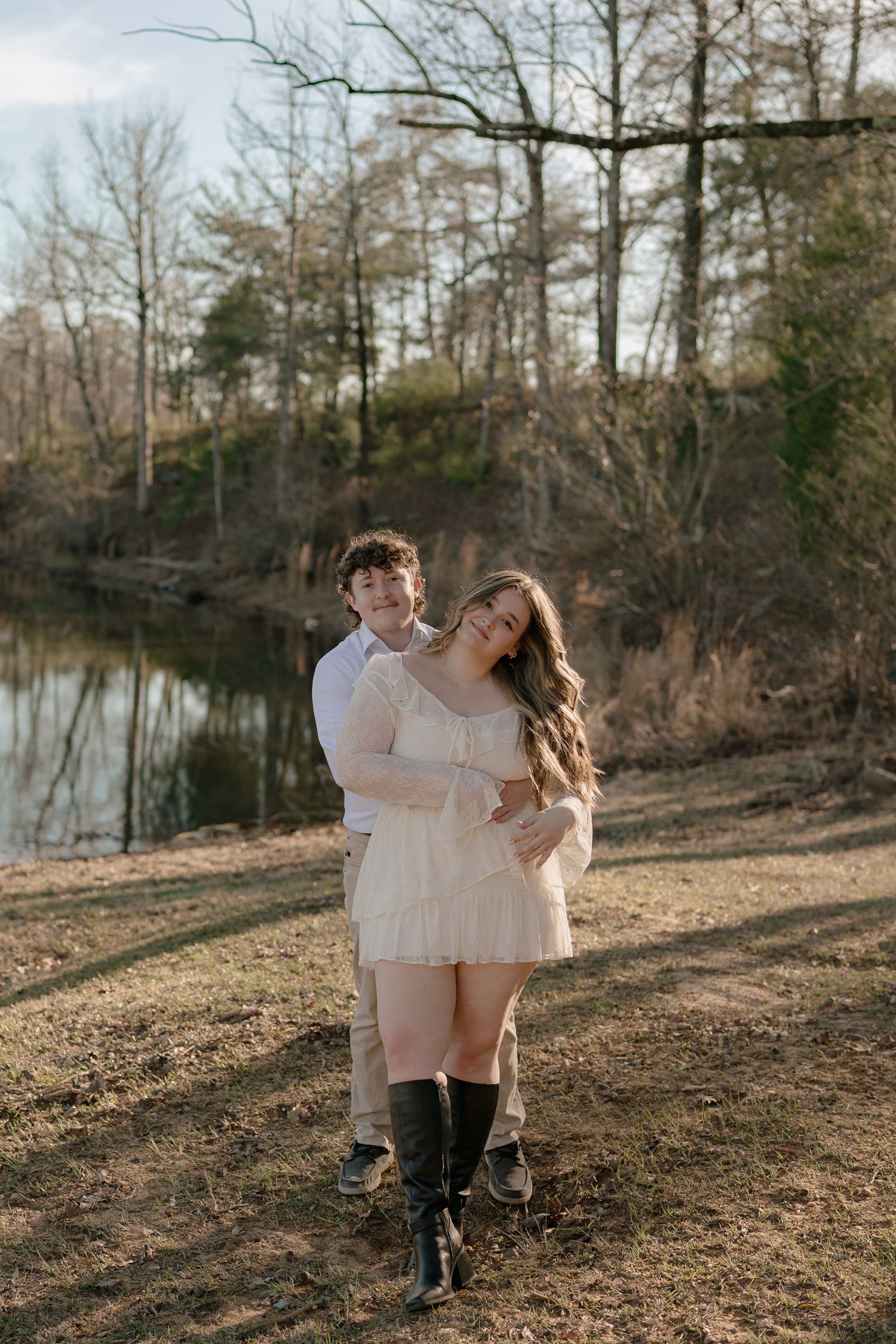 Couple embracing near a serene lake surrounded by bare trees during golden hour outdoors.