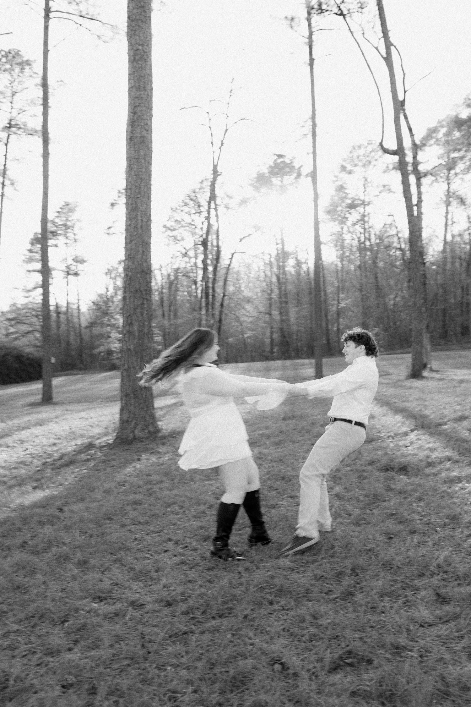 Black and white photo of a couple spinning and holding hands in a pine forest, woman in white dress and tall boots.