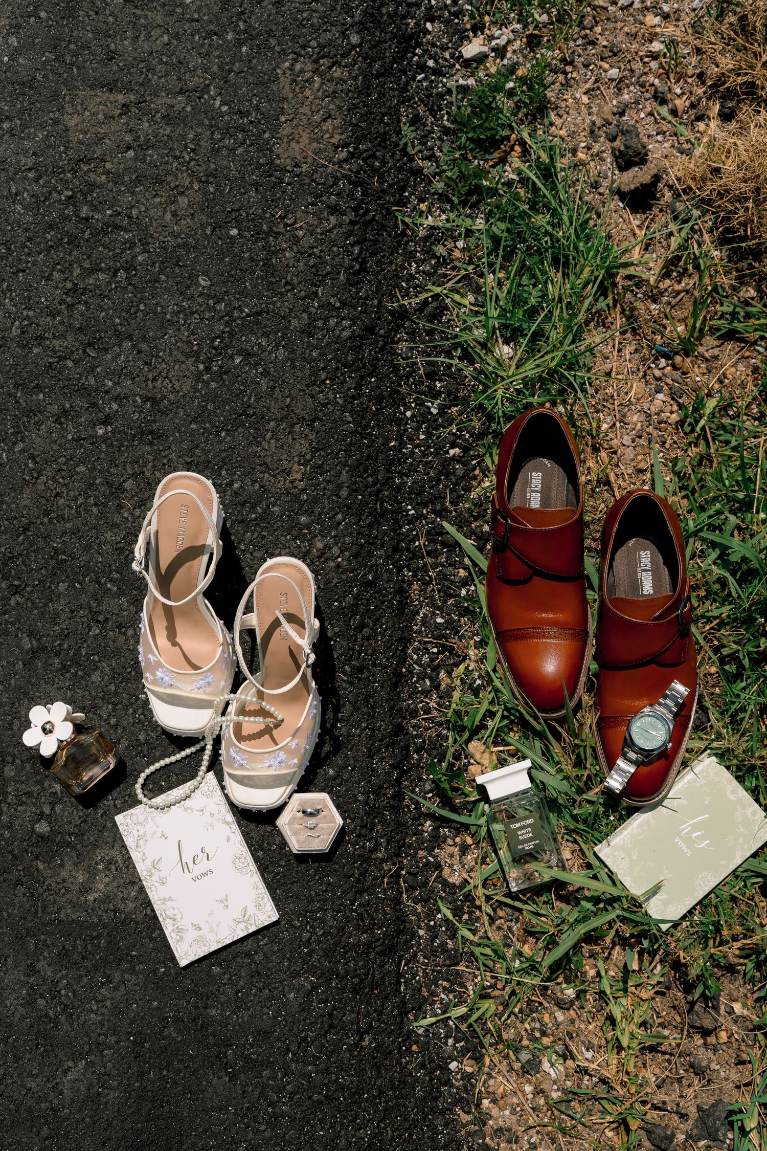 Wedding flat lay showing bride's lace heels and groom's brown leather dress shoes with accessories on pavement and grass.