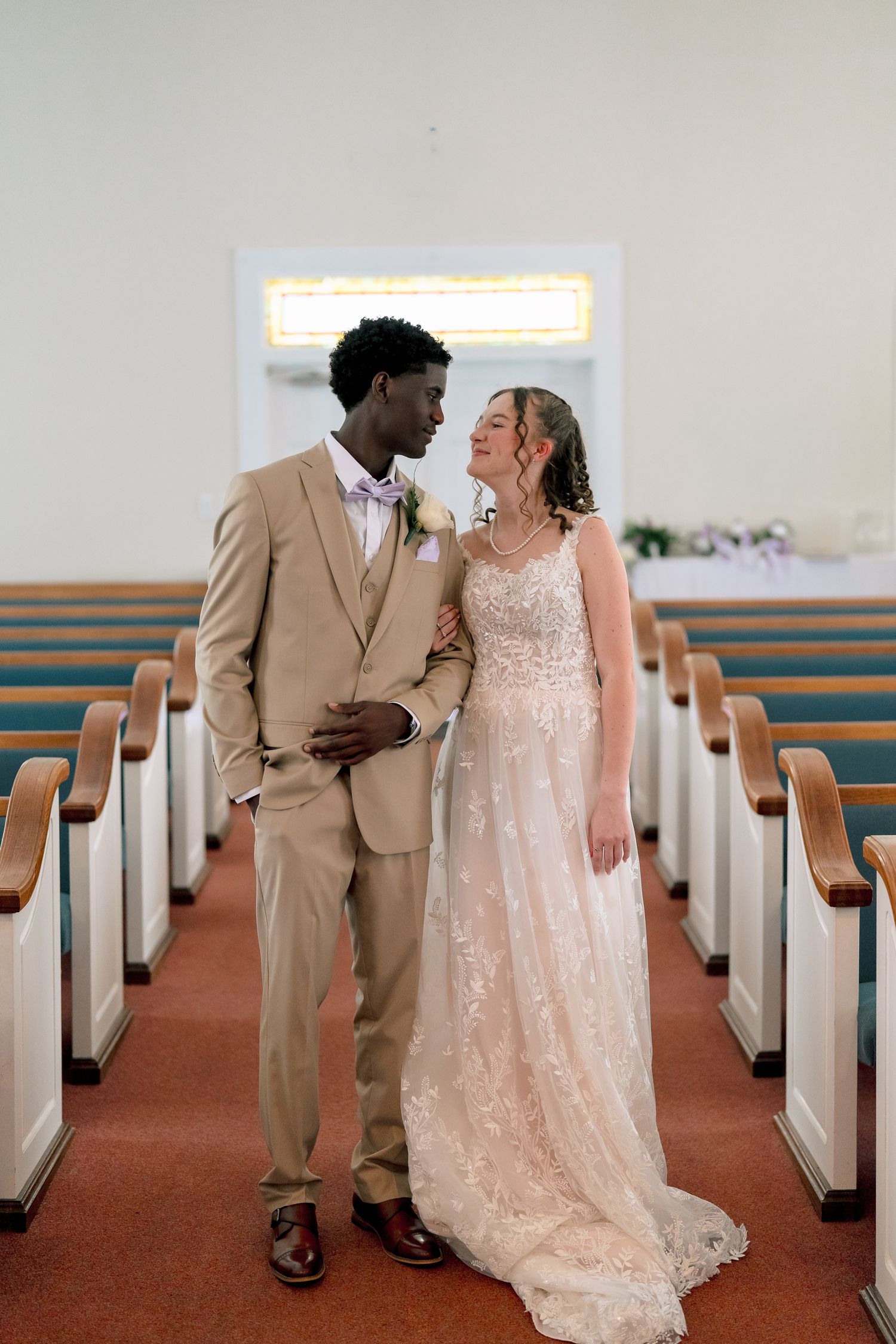 Interracial bride and groom in tan suit share romantic moment standing in church aisle between wooden pews.