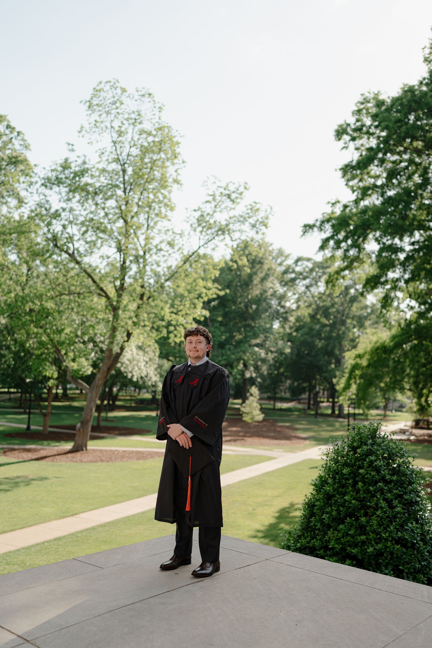 Graduate in black cap and gown stands on University of Alabama campus pathway surrounded by trees.